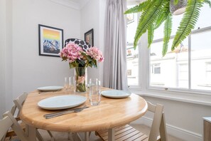 A dining area with a round wooden table, white plates, cutlery, and a vase with pink flowers. There are framed pictures on the wall and a large window with a view of buildings outside. - Host Stay Guildford Apartment (Brighton)