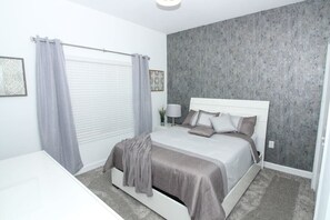 Un dormitorio moderno con una cama grande, cortinas grises, una mesita de noche blanca y una pared gris con textura.