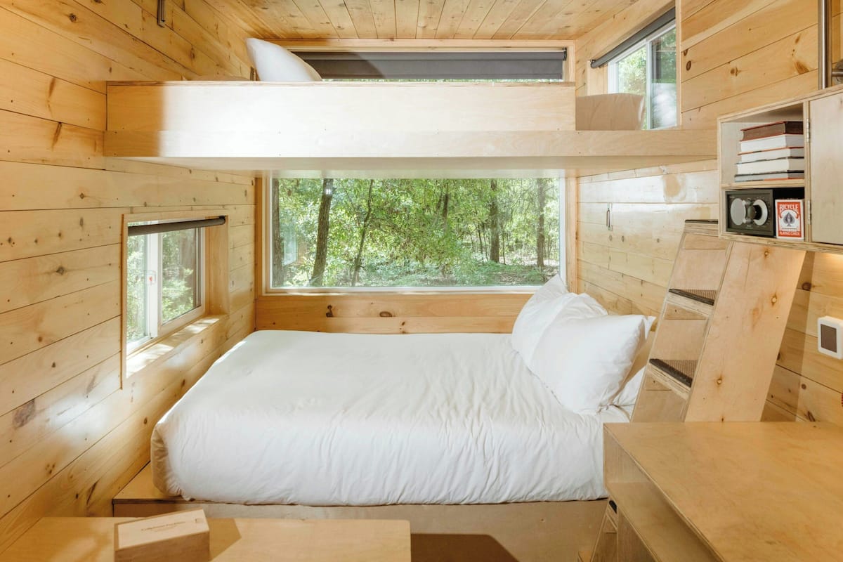 A wooden cabin bedroom with a bunk bed, large windows, and a view of the forest.
