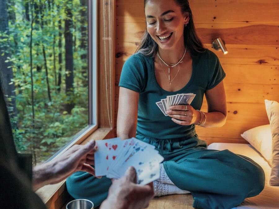 Una persona sentada en un piso de madera dentro de una cabaña, sosteniendo cartas de juego.