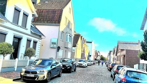 Eine gepflasterte Straße, gesäumt von parkenden Autos und Gebäuden unterschiedlicher Architektur.