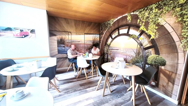 Ein gemütliches Café mit Holzboden, runden Tischen und Stühlen. Ein großes Fenster bietet Blick auf einen Strand und ein geparktes Auto.