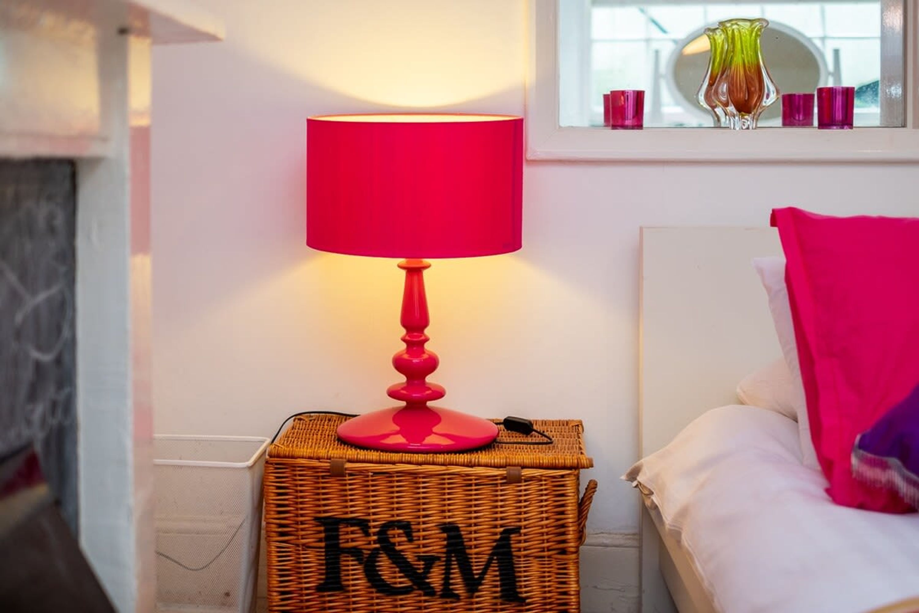 Ein Zimmer mit einer roten Lampe und einem Korbkörbchen, beschriftet mit „F&M“, neben einem Bett mit einer rosa Decke.