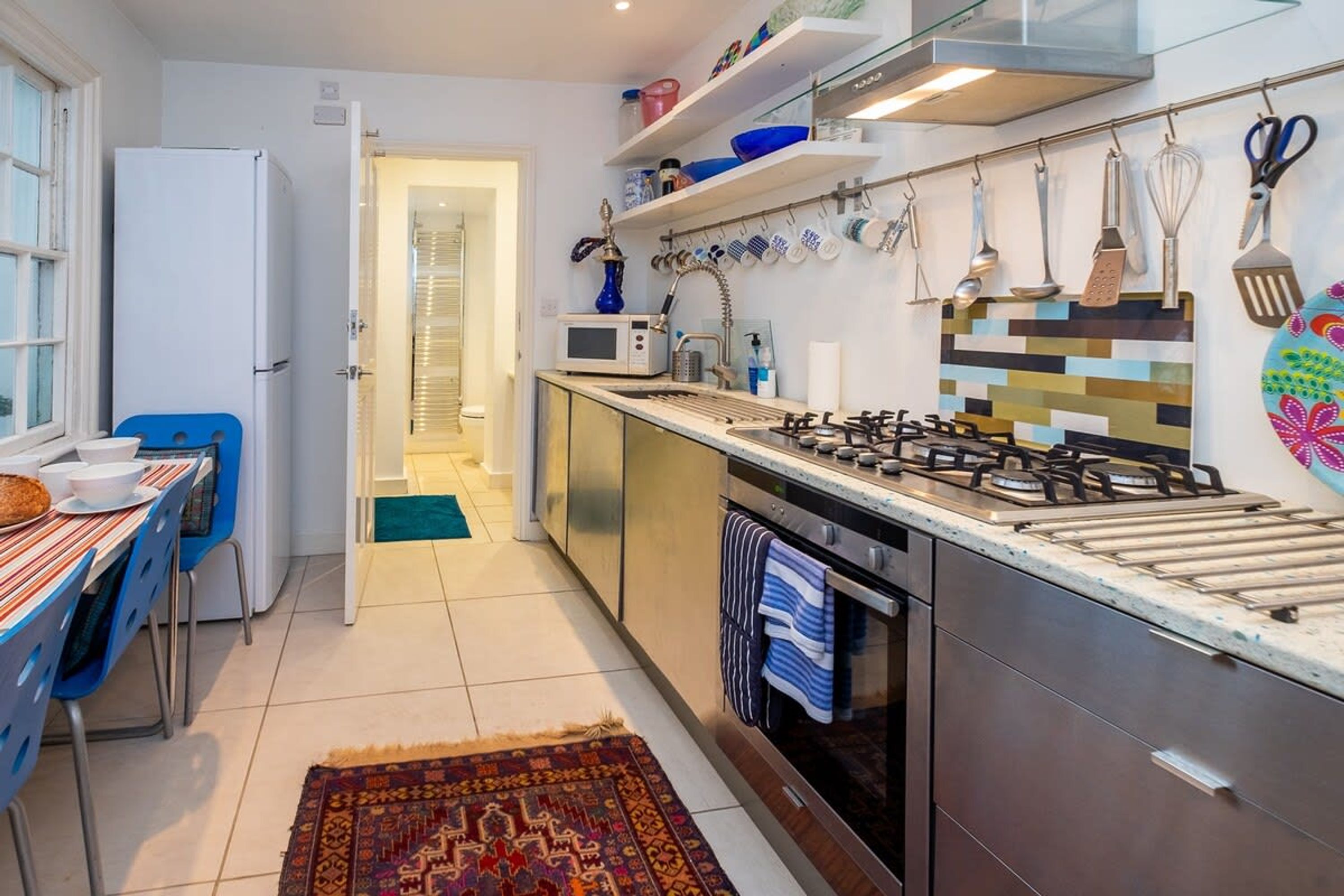 Cocina compacta con estufa a gas, microondas y armarios de acero inoxidable. Cuenta con un comedor con mesa y sillas, y una alfombra estampada en el suelo.
