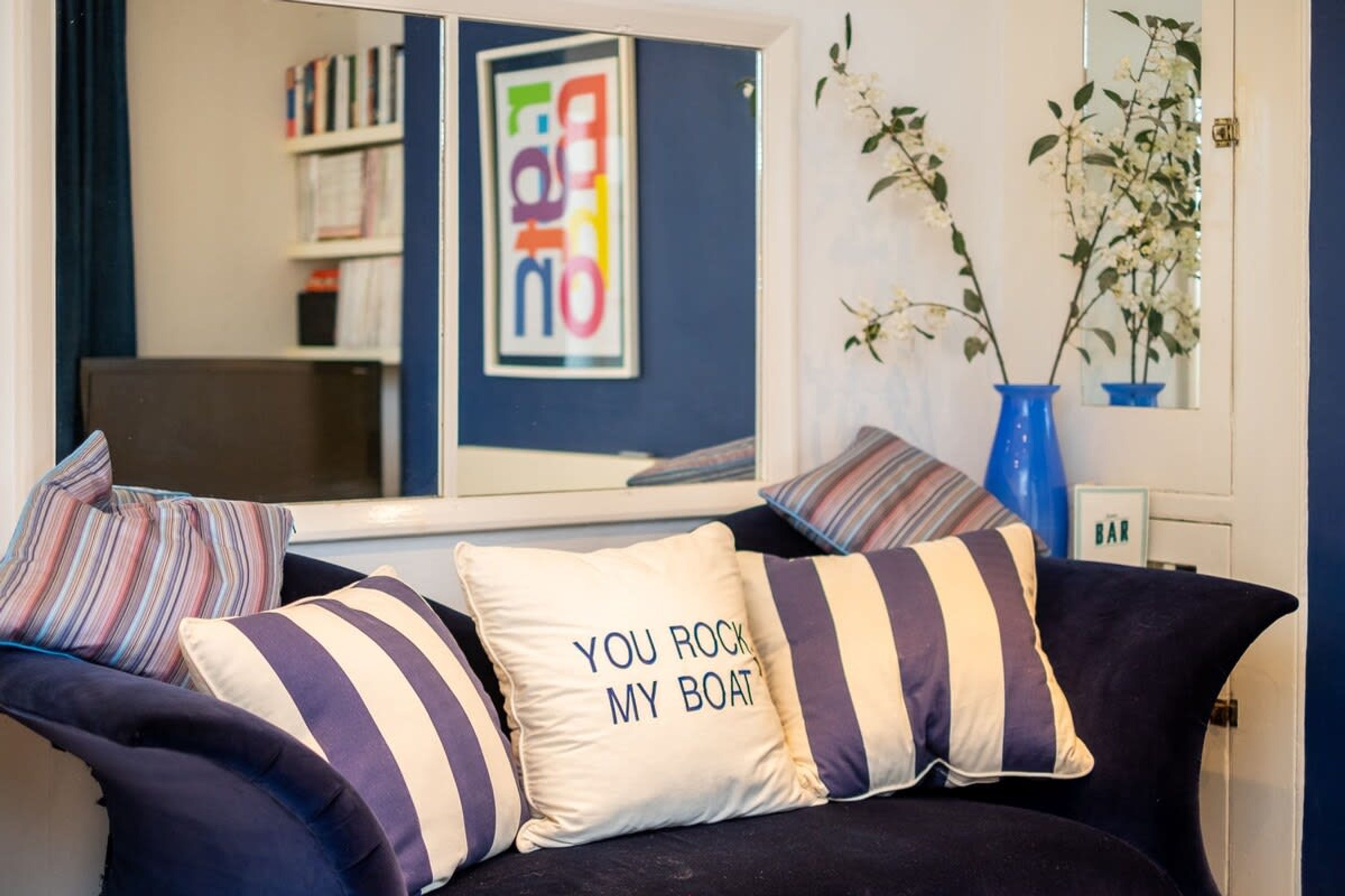 A dark blue sofa with striped pillows, a framed picture with colorful text, and a vase with white flowers.