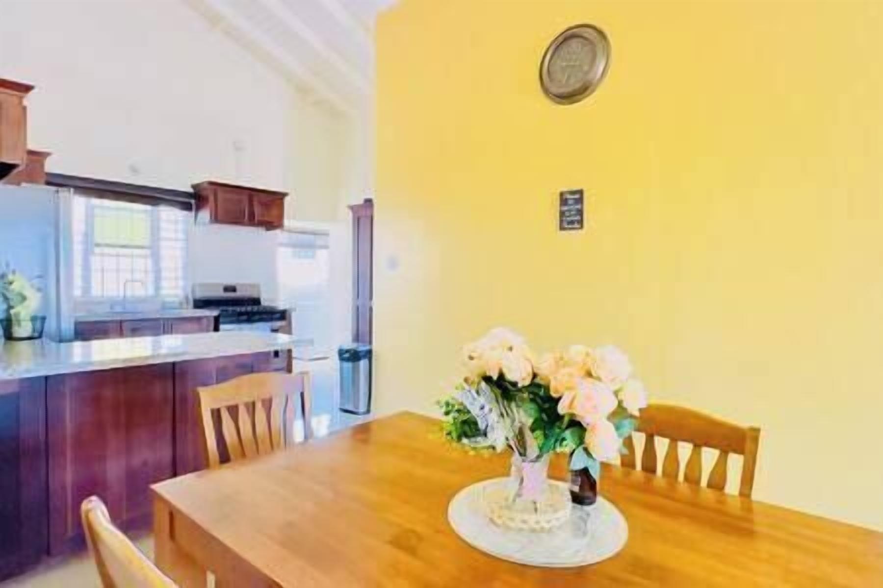 Une table à manger avec des chaises et un vase de fleurs, dans une pièce dont un mur est jaune, avec une cuisine en arrière-plan.