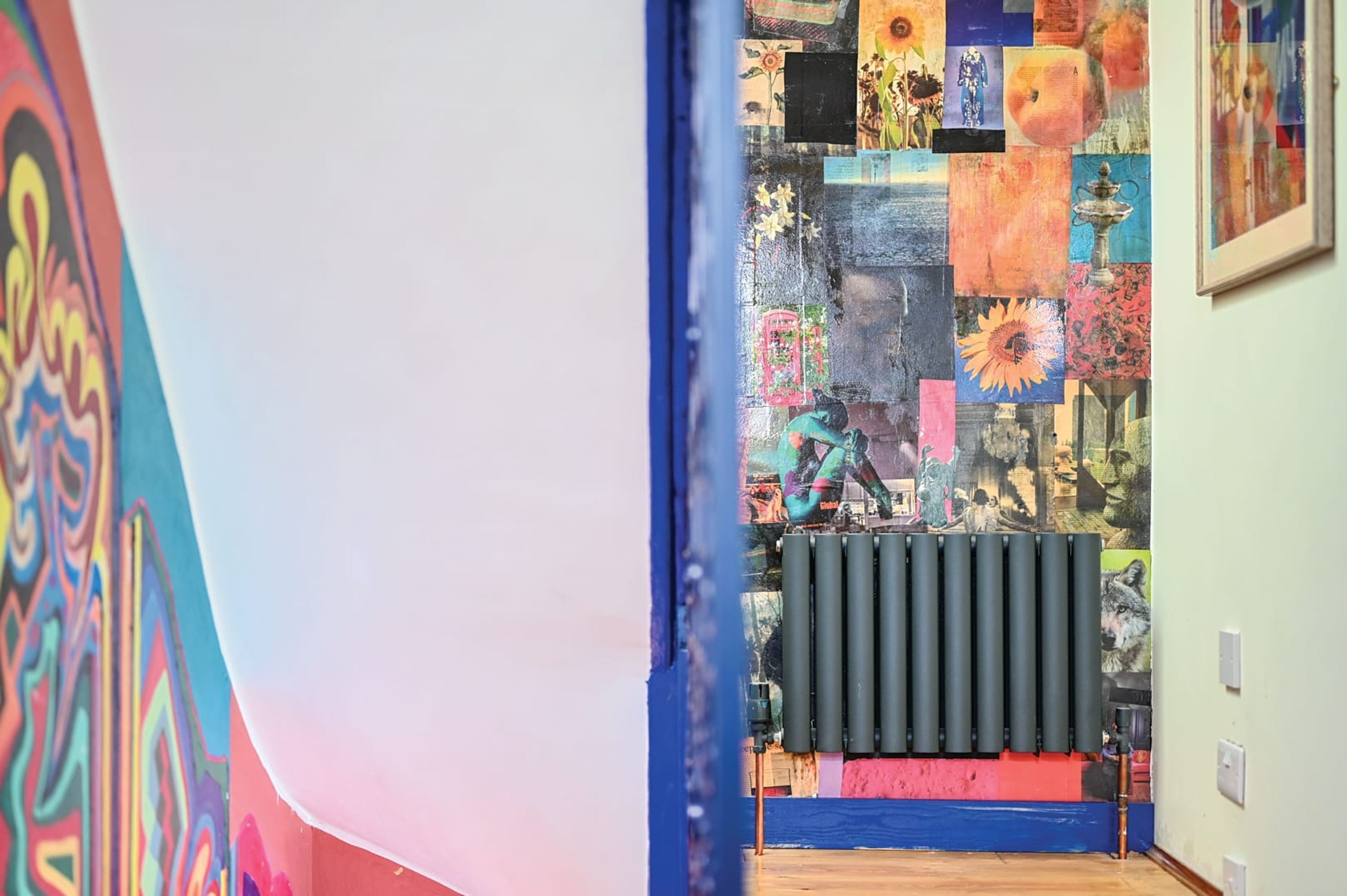 Ein Zimmer mit einem farbenfrohen, abstrakten Wandbild an der linken Wand, einem Heizkörper mit schwarzen, zylindrischen Elementen und einem Fenster mit blauem Rahmen.
