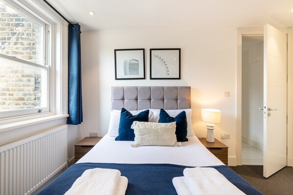 A hotel room with a bed, a window with curtains, a radiator, a bedside table with a lamp, and a door leading to another room.