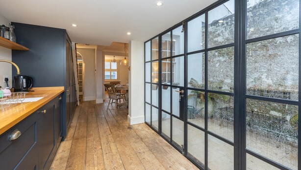 A modern kitchen with wooden flooring, dark cabinetry, and a large window with a view of the outdoors. - Host Stay The September Cottage (Brighton)