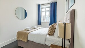 Een hotelkamer met een bed, een ronde spiegel, een raam met blauwe gordijnen, een bedlampje en een lijst met een afbeelding aan de muur.