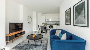 Een moderne woonkamer met een blauwe bank, een salontafel en een televisie op een houten dressoir. Er is een eethoek met stoelen en een tafel, en een keuken op de achtergrond.