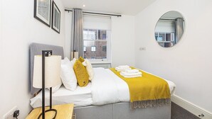 A modern hotel room with a bed, a bedside lamp, a round mirror, and framed artwork on the wall. - Modern Flat, Steps from Oxford Street! (London)
