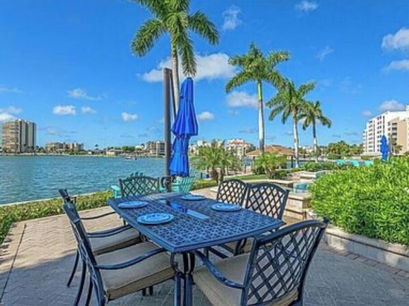 An outdoor dining area with a table and chairs, a blue umbrella, and palm trees. - Stunning Gulf waterfront pool and spa home (Marco Island)