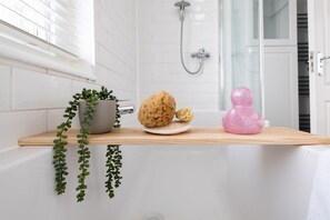 A bathroom with a white bathtub, a shower, and a wooden shelf with a potted plant, two soap bars, and a pink decorative item. - Host Stay Olde Place Cottage (Brighton)