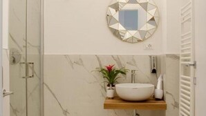 A bathroom with a marble wall, a round mirror, a wooden shelf with a white basin, a potted plant, and a towel rack. - ARENA AROUND - PRESTIGE SUITE (Verona)
