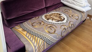 A room with a purple sofa, a decorative pillow, a stack of white towels, and a framed picture of a castle by a river. - ARENA AROUND - PRESTIGE SUITE (Verona)