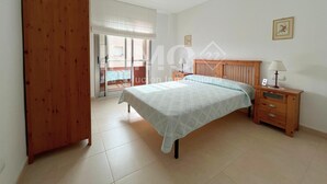 Een slaapkamer met een bed, een houten kledingkast, een nachtkastje met een lamp en een glazen schuifdeur naar een balkon.
