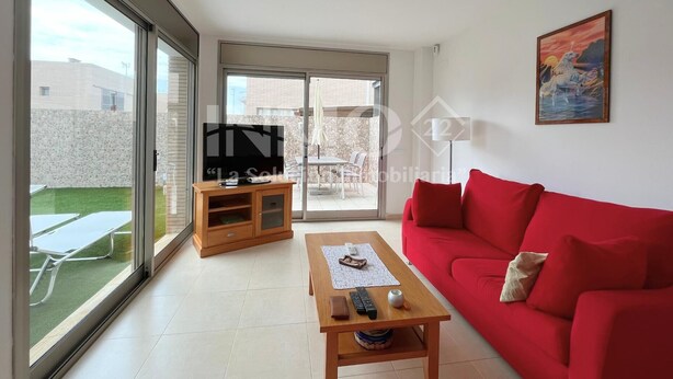 Een ruime woonkamer met een rode bank, een houten salontafel en een televisie op een kast. Een schuifdeur leidt naar een terras buiten met een tafel en stoelen.