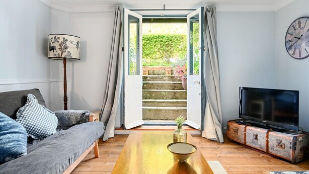 A living room with a sofa, a coffee table, a television on a vintage suitcase, and a view of a garden through open doors. - Host Stay Hanover Rest (Brighton)