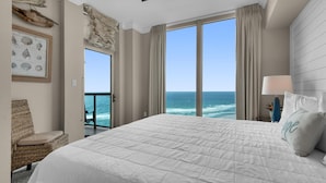 A bedroom with a large bed, a view of the ocean, a wicker chair, and a framed marine chart on the wall. - Portofino Island Resort 3-1903 (Pensacola Beach)