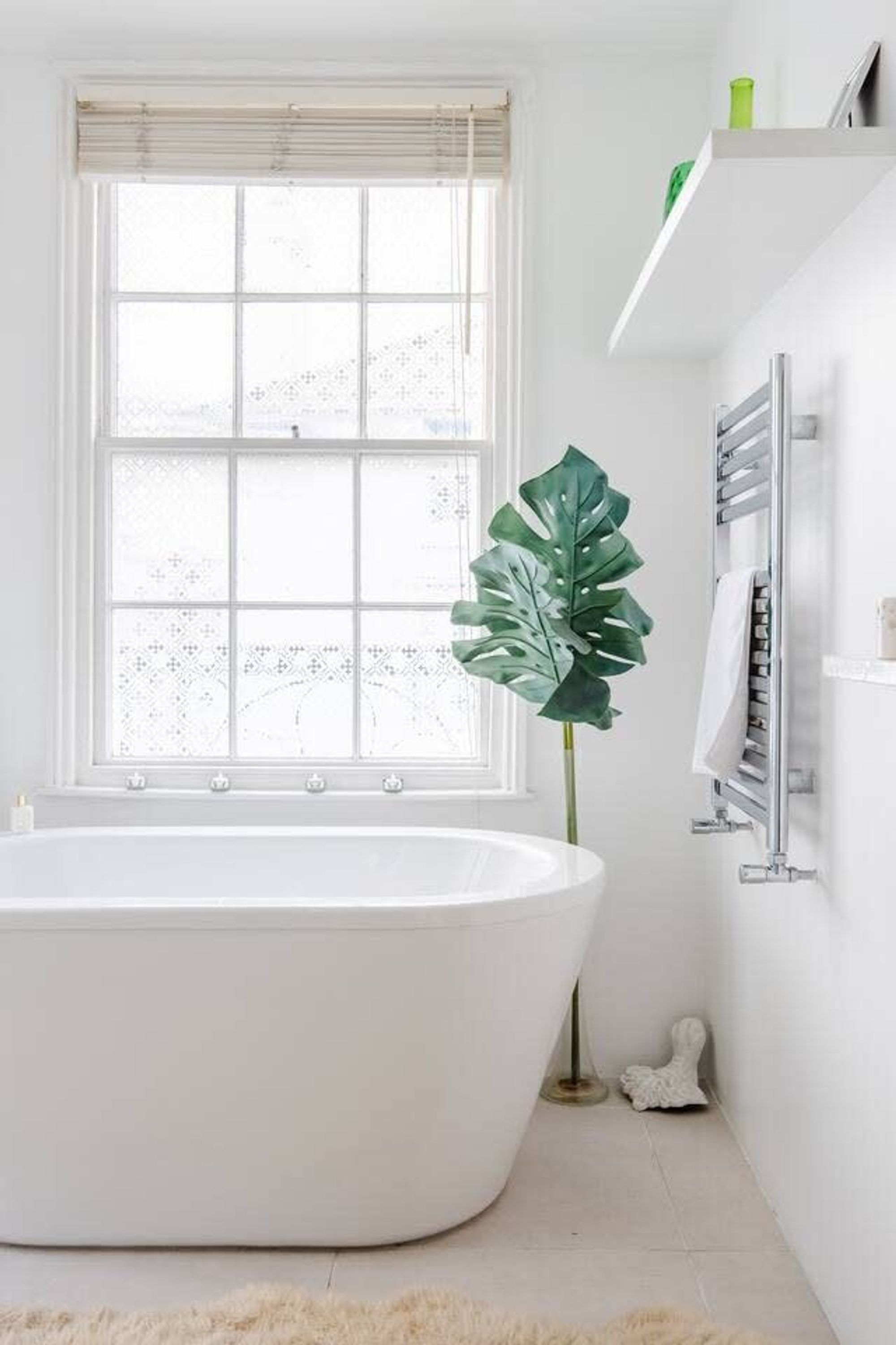 Een moderne badkamer met een vrijstaand bad, een groot raam met zonwering, een groene plant en een witte wastafel.