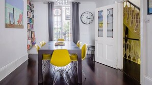 A dining area with a long table, yellow chairs, a chandelier, a clock, and a bookshelf. - Host Stay Montpelier Sanctuary (Brighton)