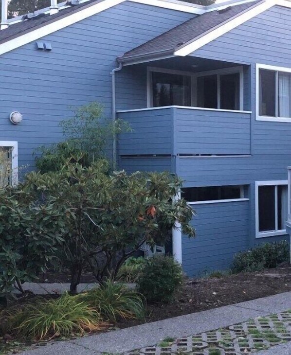 A two-story residential building with a blue exterior, a visible balcony on the upper floor, and a small garden area with various plants.