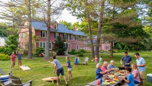 A group of people gathered around a picnic table with food, in a grassy outdoor area with trees and a building in the background. - TuckAway Suites at East Branch (Intervale)