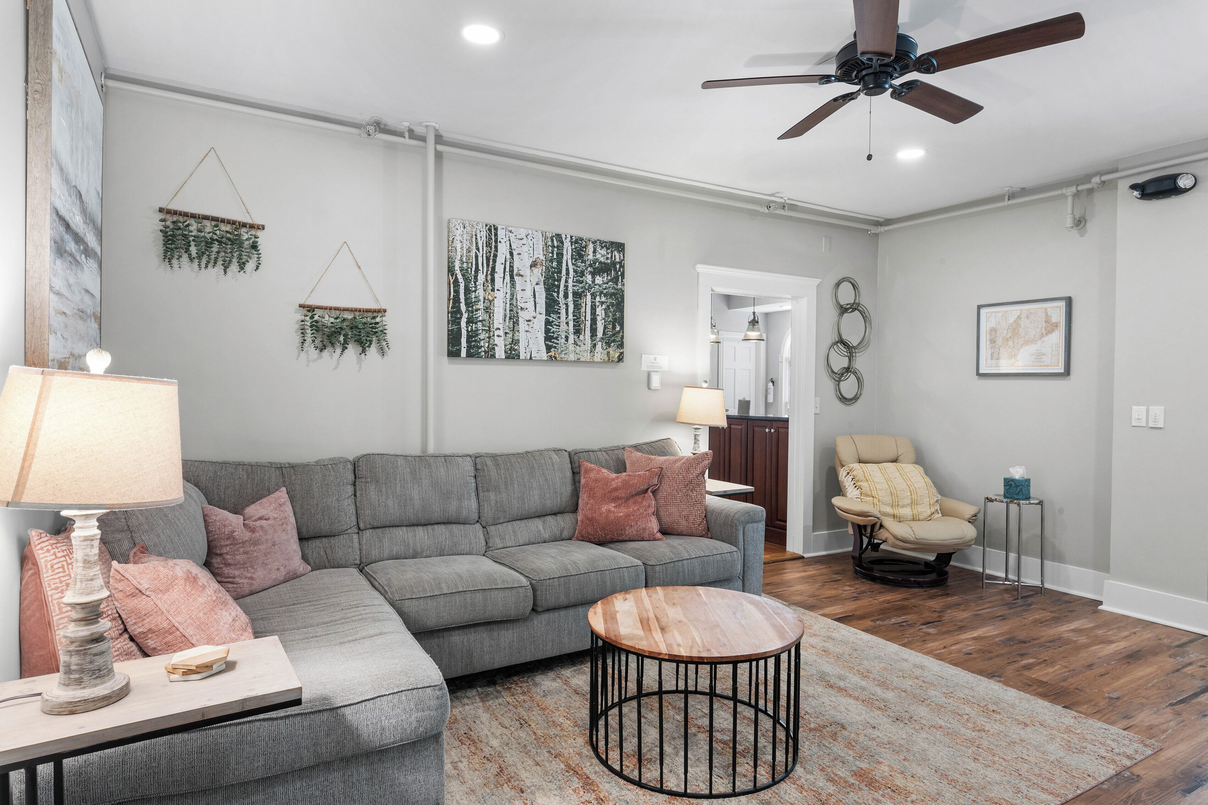 Ein Wohnzimmer mit grauem Sofa, rundem Couchtisch, Deckenventilator und Holzboden.