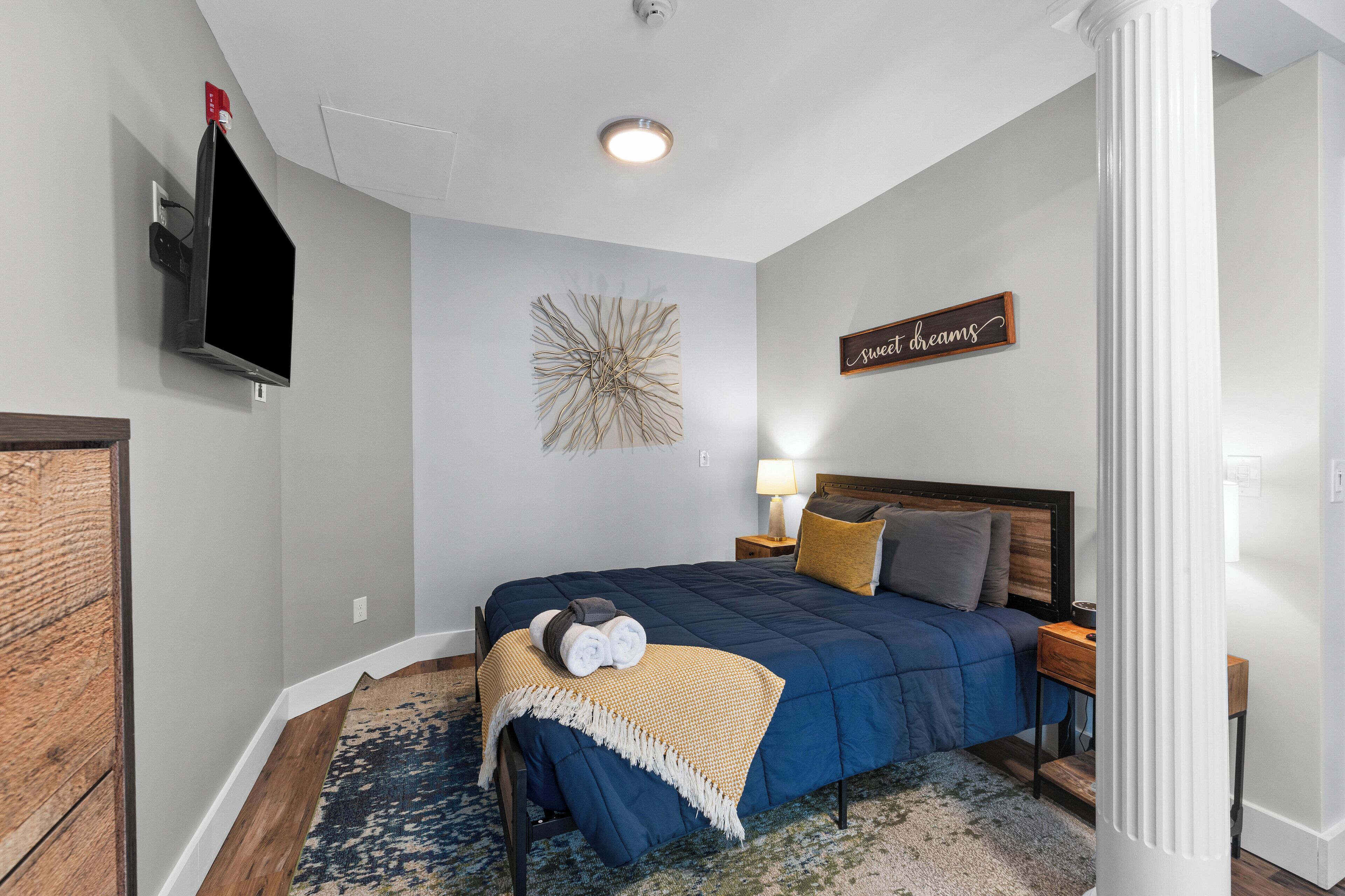 Un dormitorio con cama, televisor montado en la pared, adorno decorativo en la pared, lámpara de noche y un cartel que dice 'Sweet dreams'.