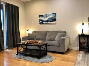 A living room with a grey sofa, a coffee table, a painting on the wall, a bookshelf, and a window with curtains.