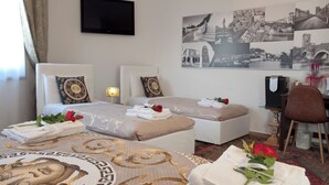 A hotel room with two beds, a flat-screen TV, a desk with a chair, and a wall with black and white photographs. - ARENA AROUND - IMPERIAL SUITE (Verona)