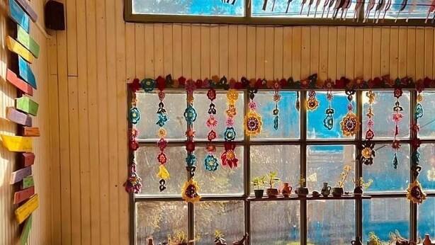 A cozy room with a wooden wall, a window with colorful decorations, a couch, and a bookshelf. - Casa Piwke (Coyhaique)