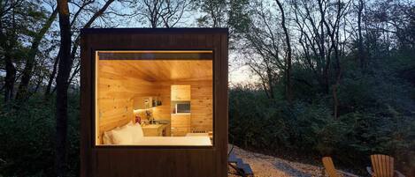 A wooden cabin with a kitchen and a bed, surrounded by trees and a fire pit.