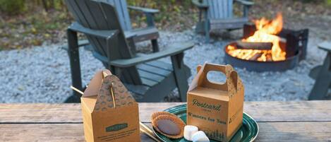 A picnic setup with Adirondack chairs, a campfire, and two paper bags labeled "S'mores for Two" on a wooden table.