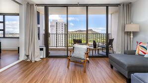 A spacious room with a balcony, wooden flooring, a grey sofa, a chair, and a table with a lamp.