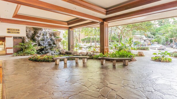 A spacious lobby with a stone floor, benches, a waterfall feature, and a sign for 'Tower 1 Makai'.