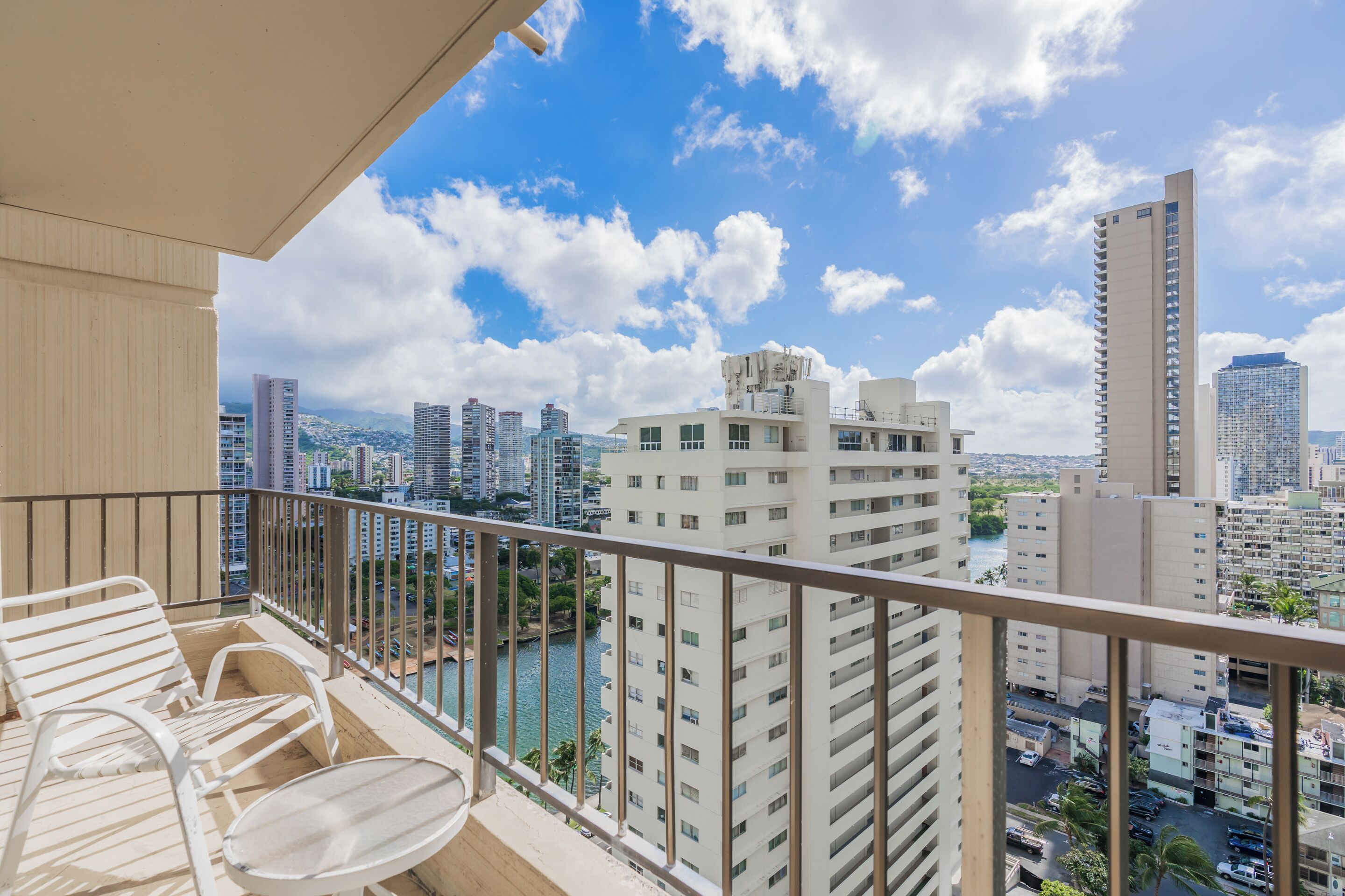 A balcony with white chairs and a table, overlooking a cityscape with tall buildings and a river.