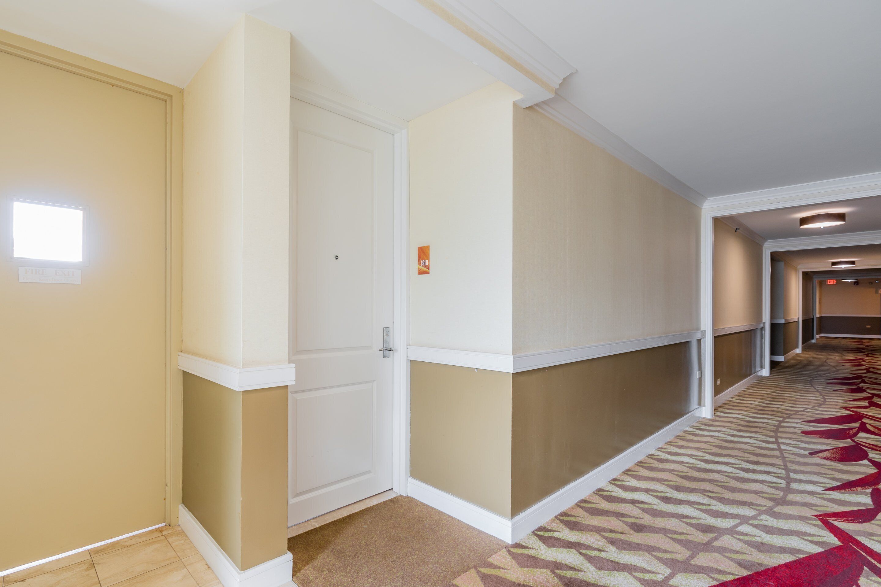 A hotel hallway with beige walls, a patterned carpet, and a white door.