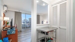 A hotel room with a wooden desk, a chair, a lamp, a television, and a view of the city. There is a metal shelf with kitchenware and a white closet with louvered doors. - 🍧 Summer Special - Royal Garden Waikiki - Ocean View - Resort Amenities 😎 (Honolulu)