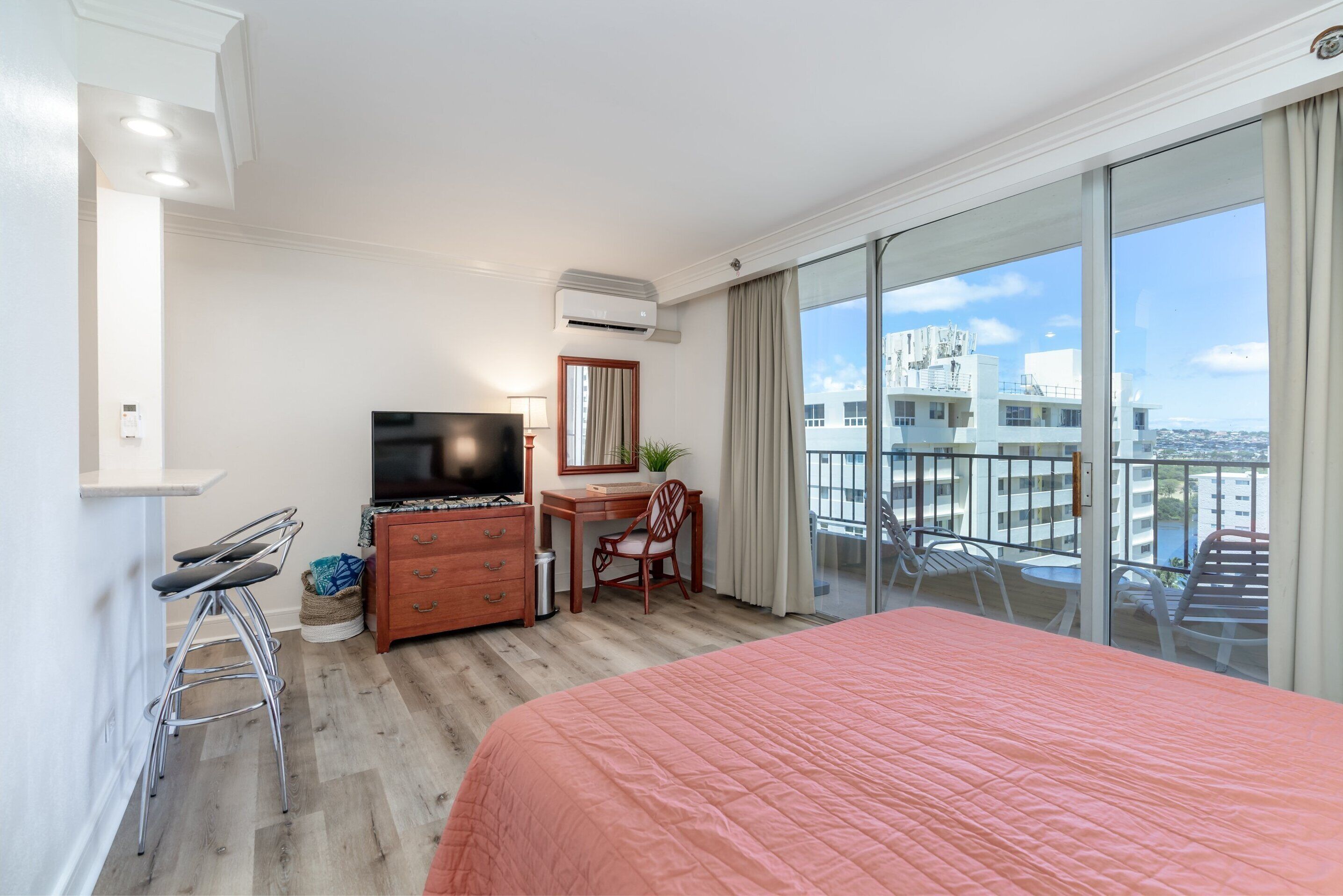 A hotel room with a bed, a desk, a chair, a TV, and a balcony with a view of buildings.