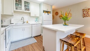 A modern kitchen with white cabinets, a built-in dishwasher, and a central island with stools. - Hale Kai - Large Enclosed Lanai - Sleeps 2 (Kahuku)