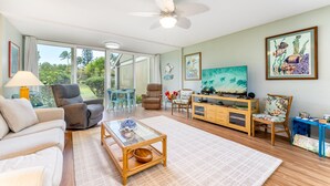 A spacious living room with a sofa, chairs, a coffee table, and a television. There is a ceiling fan, a large window with a view of greenery, and a painting on the wall. - Hale Kai - Large Enclosed Lanai - Sleeps 2 (Kahuku)