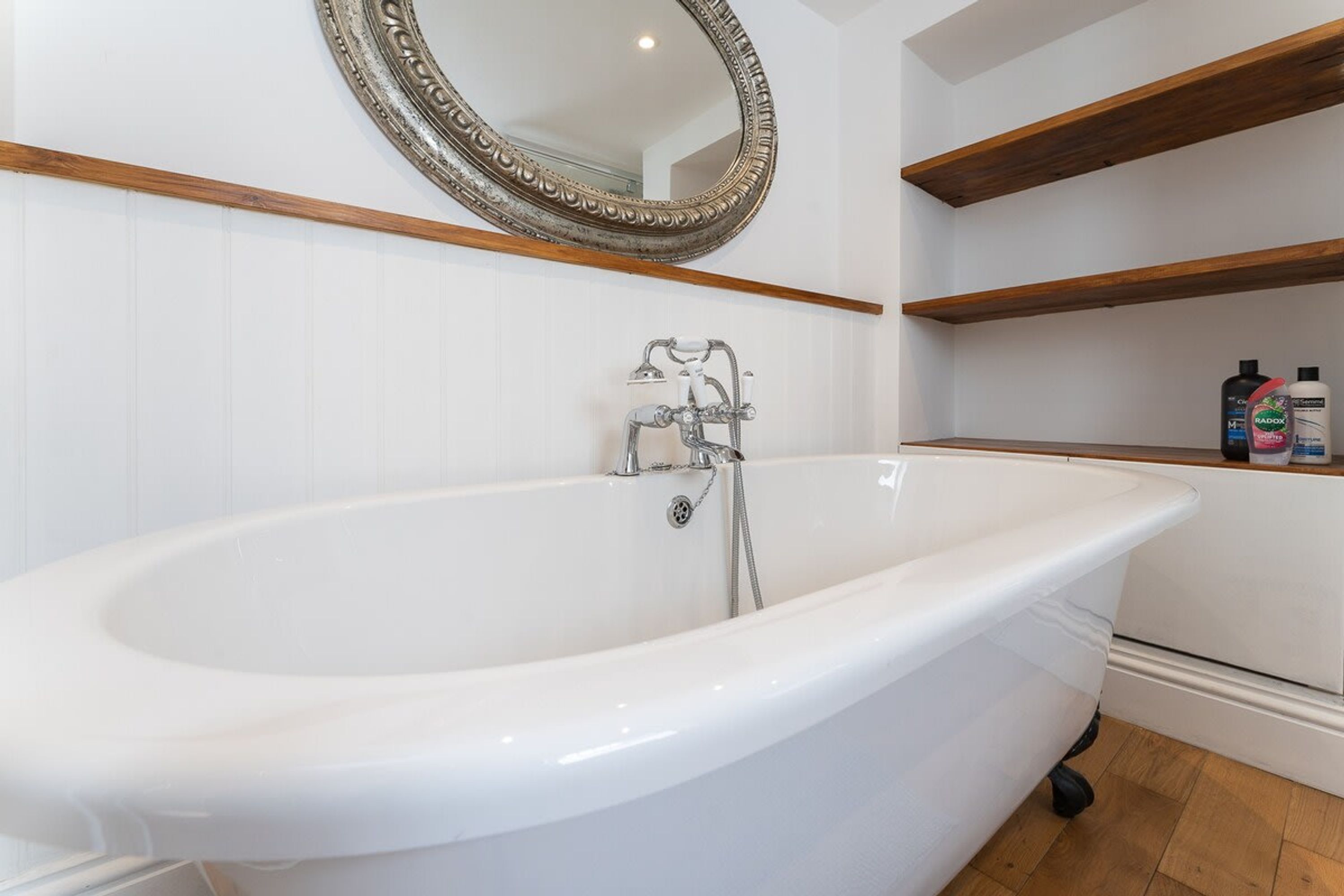 A white freestanding bathtub with a modern faucet, a wooden shelf above, and a mirror with an ornate frame.