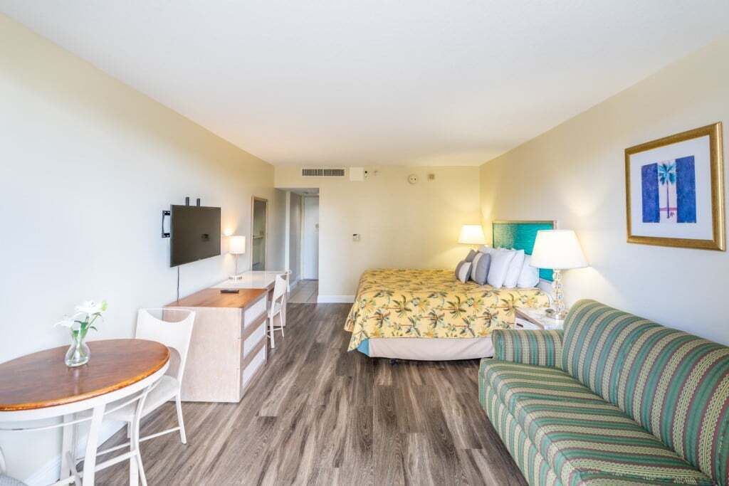 A hotel room with a bed, a sofa, a desk with a chair, a small table, and a TV mounted on the wall.