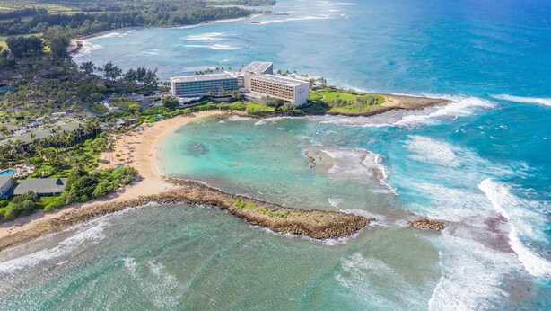 An aerial view of a coastal resort with a large building, a sandy beach, and clear turquoise waters. - 😎🍧Summer Special - The Drifter - North Shore Loft - Relaxed Luxury ☀️🏄‍♀️ (Kahuku)