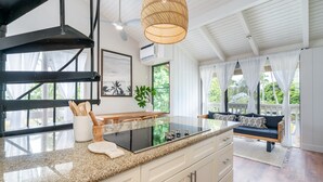 A modern kitchen with a granite countertop, a built-in stove, and white cabinetry. There is a dining area with a bench and a hanging light fixture. - 😎🍧Summer Special - The Drifter - North Shore Loft - Relaxed Luxury ☀️🏄‍♀️ (Kahuku)