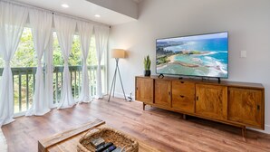 A spacious living room with a large flat-screen TV, wooden flooring, and a view of a beach through large windows. - 😎🍧Summer Special - The Drifter - North Shore Loft - Relaxed Luxury ☀️🏄‍♀️ (Kahuku)