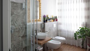 Une salle de bain moderne avec une douche à parois de verre, un grand miroir avec un cadre doré, un lavabo blanc, un bidet et une plante en pot.