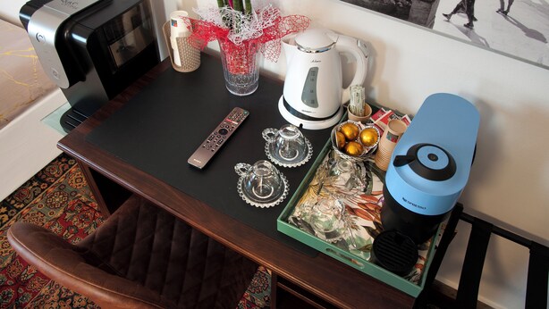 Une chambre d’hôtel avec une table à manger, une machine à café, une théière, des tasses et un plateau de fruits. On peut également apercevoir une télécommande et une chaise.